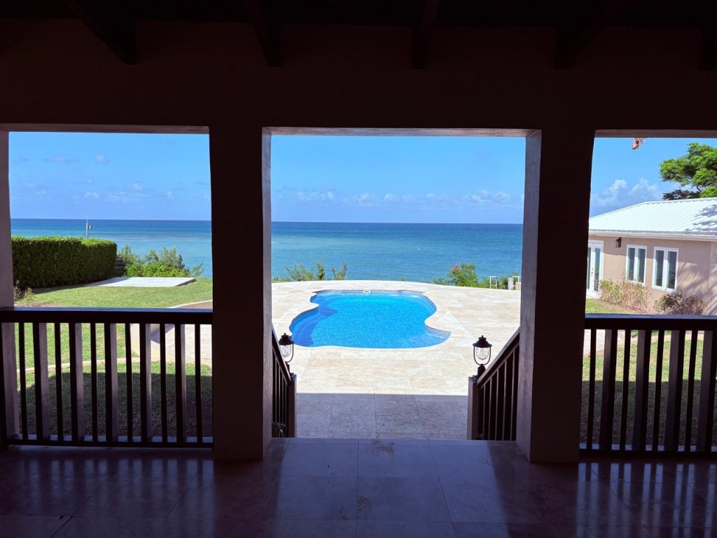 A serene view of a pool and the ocean from a covered porch with a railing.
